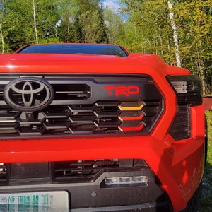 May include: A red Toyota Tacoma pickup truck with a black grille and a red "TRD" emblem. The grille has a honeycomb pattern and a black Toyota logo. The truck has a black license plate with the text "LIVING FREE TO DIE".