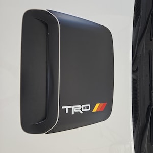 May include: A matte black hood scoop mounted on a white vehicle surface. The scoop features a prominent air intake vent on the left side and the white text TRD followed by three slanted stripes in yellow, orange, and red on the lower right corner.