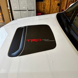 May include: White car hood with a black and gray graphic of a topographic map. The graphic includes the red text "TRD OFF ROAD".