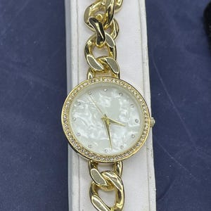 May include: Gold-toned wristwatch with a chain-link band. The watch face is round, with a white mother-of-pearl effect and a ring of small crystals. The hands are gold, and the band is a chunky, polished gold.