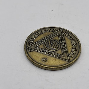 May include: A bronze-colored coin with the words "TO THINE OWN SELF BE TRUE" around the edge. The center features a triangle with "UNITY SERVICE RECOVERY" and the Roman numeral XIII.