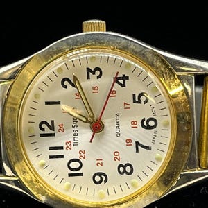 Puede incluir: Un reloj de pulsera dorado y plateado con esfera crema. La esfera del reloj presenta números negros, detalles en rojo y el texto "Times Square" y "Quartz". La correa del reloj es una combinación de eslabones dorados y plateados.