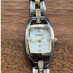 Puede incluir: Un reloj Fossil de tono plateado y dorado con una esfera blanca y detalles de diamantes en la correa. La esfera del reloj tiene un efecto de nácar y la palabra "Fossil" está impresa en la esfera.