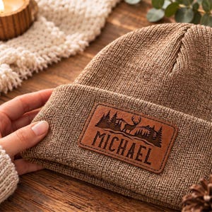 May include: A brown knit beanie featuring a rectangular leather patch on the cuff. The patch is engraved with a forest landscape, a deer silhouette, and the name MICHAEL in capital letters centered below the graphic design.