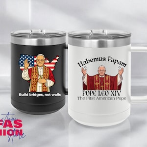 May include: Two travel mugs, one black and one white, with silver lids and handles. The black mug features an image of Pope with the American flag and the text "Build bridges, not walls." The white mug has the text "Habemus Papam" and "Pope Leo XIV, The First American Pope."