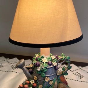 May include: A decorative lamp featuring a watering can base adorned with blackberries, flowers, and a rabbit. The lamp has a beige lampshade with a black trim. The base includes a small pot of strawberries and a garden trowel.