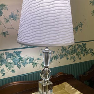 Puede incluir: Una lámpara de mesa de cristal transparente con una pantalla blanca de textura ondulada. La lámpara tiene una base geométrica y una columna decorativa de cristal. La lámpara está sobre una mesa con una tela dorada.