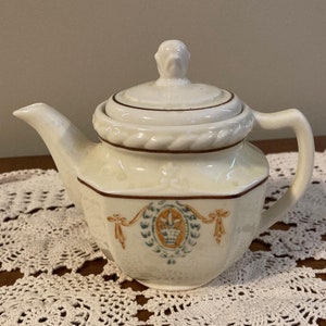 May include: A white porcelain teapot with a lid and a floral design. The teapot has a brown trim and a gold accent. The teapot is sitting on a white lace tablecloth.