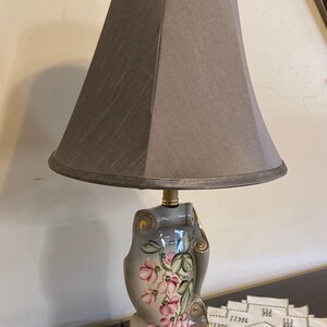 May include: A decorative table lamp with a gray lampshade and a floral-painted base. The base features pink flowers and gold accents. The lamp has a brass finial and a marble-like base, adding a touch of elegance to any room.