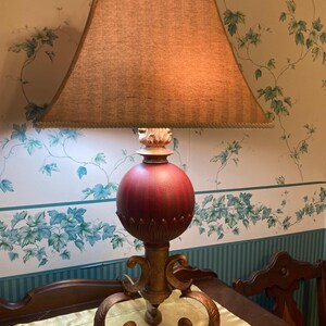 May include: A decorative table lamp with a brown lampshade and a red and gold base. The lamp has a square lampshade and a detailed base with a spherical red center. The lamp is on a table with a gold tablecloth.