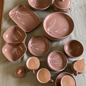 Könnte beinhalten: Eine Sammlung von Vintage-Geschirr aus brauner Keramik mit weißem Blattdesign. Das Set umfasst Teller, Schalen, Tassen, Untertassen, einen Sahnekännchen und eine Schüssel mit Deckel. Das Geschirr ist auf einer hellgrünen Oberfläche angeordnet.
