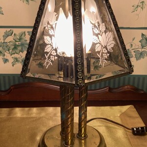 May include: Vintage brass table lamp with a clear glass shade featuring a floral design. The lamp has a round base and two twisted support columns. The shade is a trapezoid shape. The lamp is turned on, illuminating the room.