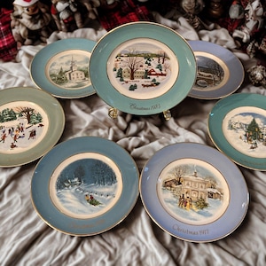 May include: A collection of vintage decorative plates with winter scenes. Each plate features a different illustration of a snowy landscape with buildings and people. The plates have a gold rim and are labeled with the year "Christmas".