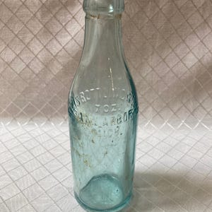Peut inclure: Une bouteille en verre bleu clair vintage avec du texte en relief. La bouteille a un col étroit et une base arrondie. Le texte indique "H.S. Scott-Work" et "7 OZ. ANN ARBOR MICH." La bouteille est légèrement sale, montrant son âge.