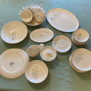 May include: A collection of vintage white ceramic dishware with a yellow floral design. The set includes plates, bowls, cups, saucers, a serving platter, a creamer, and a sugar bowl. The dishes have a gold trim.