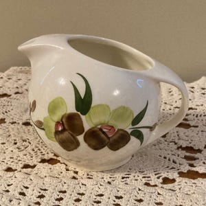 May include: White ceramic pitcher with a curved handle and spout. The pitcher is decorated with hand-painted flowers in shades of brown, green, and yellow. The pitcher sits on a cream-colored lace tablecloth.