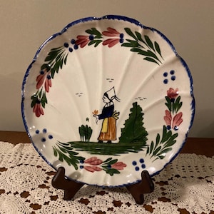 May include: A white ceramic plate with a scalloped edge, featuring a hand-painted design of a woman in a blue dress and white hat holding a flower. The plate is decorated with green leaves, pink flowers, and blue dots.