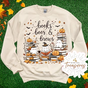 May include: Cream-colored sweatshirt with a Halloween-themed graphic. The design features stacks of books, pumpkins, ghosts, bats, and the text "books, boos & brews". The graphic is in shades of orange, brown, and black.