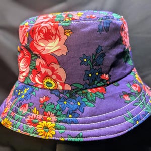May include: A purple bucket hat with a floral print featuring pink, red, yellow, blue, and green flowers. The hat has a wide brim and white stitching detail.