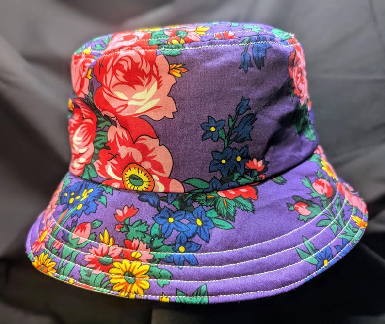 May include: A purple bucket hat with a floral print featuring pink, red, yellow, blue, and green flowers. The hat has a wide brim and white stitching detail.