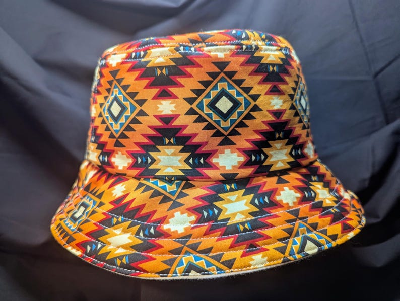May include: A bucket hat featuring a geometric pattern in shades of orange, red, yellow, blue, and black. The pattern is reminiscent of Native American designs.