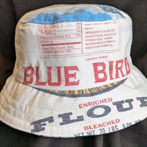 May include: A white bucket hat featuring a blue bird design and the words "Blue Bird Enriched Flour Bleached Net Wt 20 Lbs 9.08 Kg" printed on it. The hat is made from recycled flour sacks.