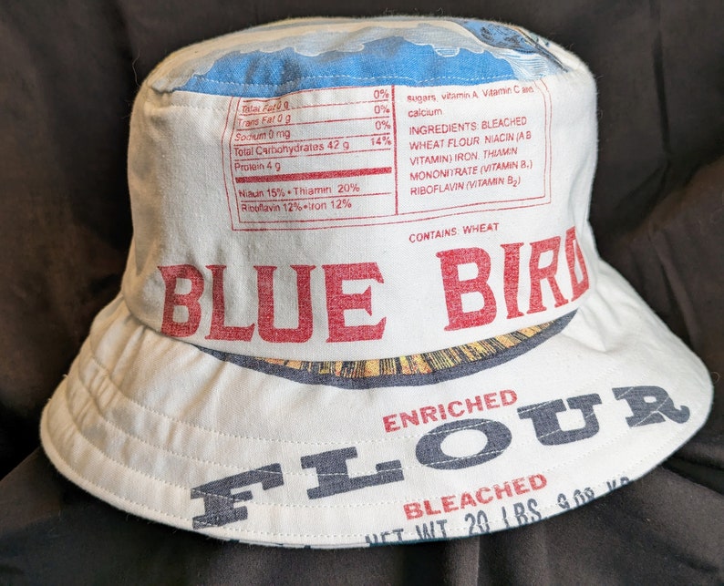 May include: A white bucket hat featuring a blue bird design and the words "Blue Bird Enriched Flour Bleached Net Wt 20 Lbs 9.08 Kg" printed on it. The hat is made from recycled flour sacks.