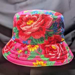May include: A pink bucket hat with a floral print featuring red, pink, yellow, blue, and green flowers. The hat has a wide brim and a flat crown.