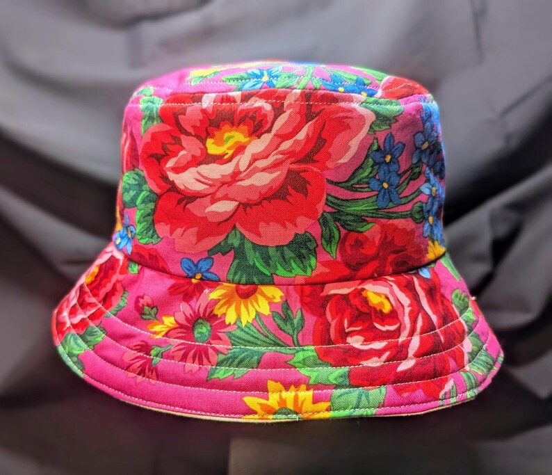 May include: A pink bucket hat with a floral print featuring red, pink, yellow, blue, and green flowers. The hat has a wide brim and a flat crown.