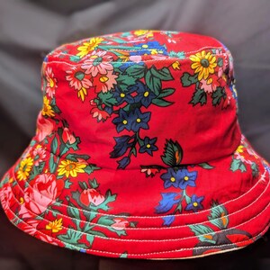 May include: A red bucket hat with a floral print featuring yellow, blue, green, and pink flowers. The hat has a wide brim and a white lining.