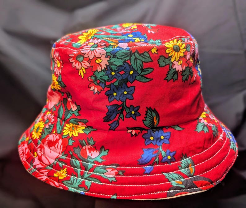 May include: A red bucket hat with a floral print featuring yellow, blue, green, and pink flowers. The hat has a wide brim and a white lining.