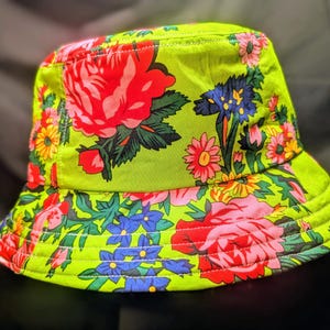 May include: A green bucket hat with a floral print featuring pink, red, blue, and yellow flowers. The hat has a wide brim and white stitching detail.