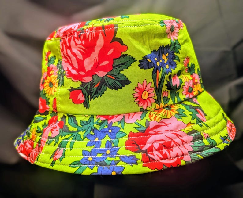 May include: A green bucket hat with a floral print featuring pink, red, blue, and yellow flowers. The hat has a wide brim and white stitching detail.