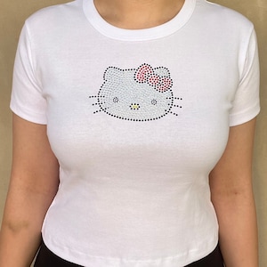 Pu&ograve; includere: Maglietta bianca corta con un design Hello Kitty in strass. Il design include un fiocco rosso e baffi neri.