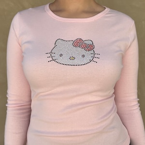 May include: A pink long-sleeved shirt with a rhinestone Hello Kitty design. The design features a white Hello Kitty face with a red bow.