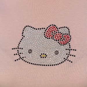 May include: A pink fabric with a rhinestone Hello Kitty design. The design features a white Hello Kitty face with a red bow and black outlines.
