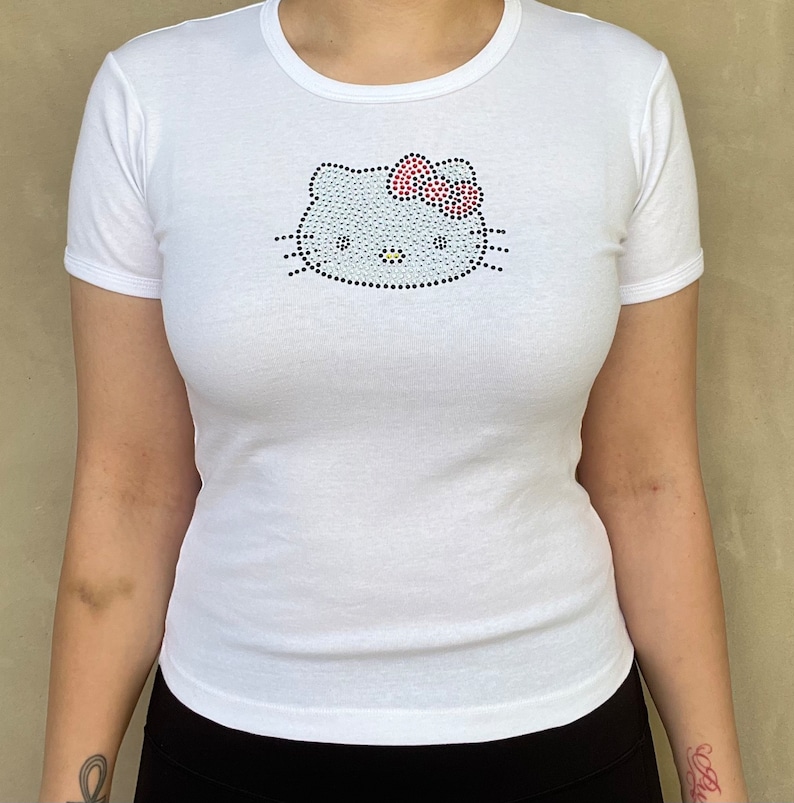 Puede incluir: Camiseta blanca con un dise&ntilde;o de Hello Kitty de pedrer&iacute;a. El dise&ntilde;o presenta una cara de Hello Kitty blanca con un lazo rojo.