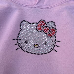 Puede incluir: Una sudadera con capucha de color morado claro con un dise&ntilde;o de Hello Kitty de diamantes de imitaci&oacute;n. El dise&ntilde;o tiene un contorno negro y un lazo rojo.