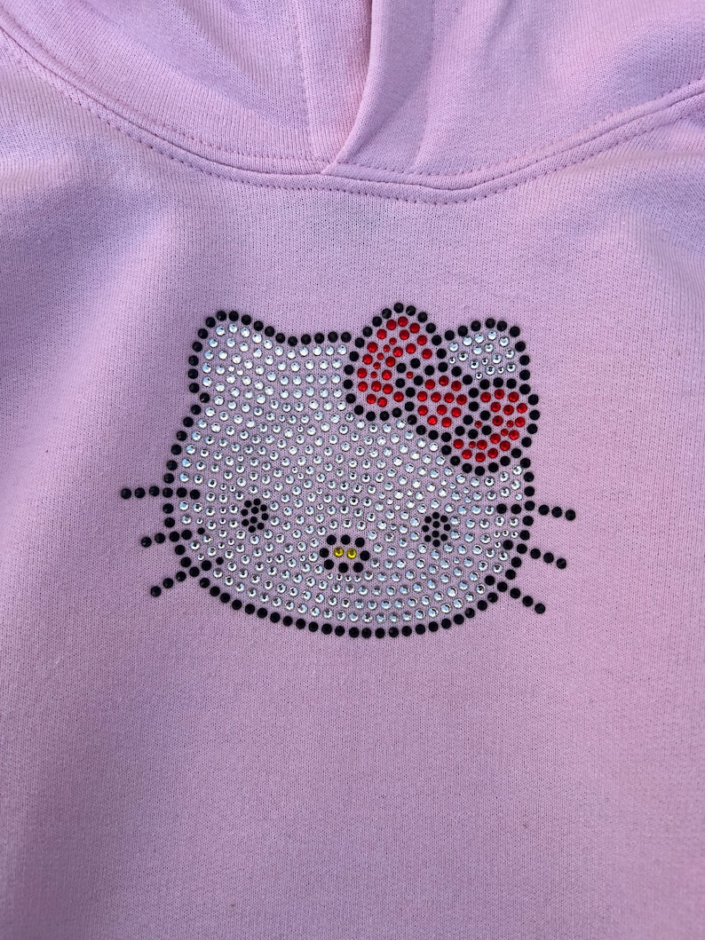 Puede incluir: Una sudadera con capucha de color morado claro con un dise&ntilde;o de Hello Kitty de diamantes de imitaci&oacute;n. El dise&ntilde;o tiene un contorno negro y un lazo rojo.