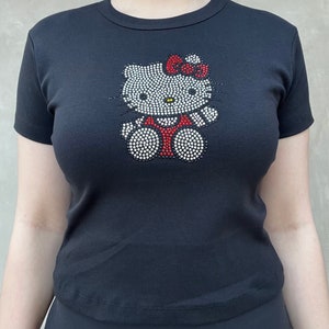 May include: Black t-shirt featuring a rhinestone Hello Kitty design. The design has a white outline, red bow, and black eyes.