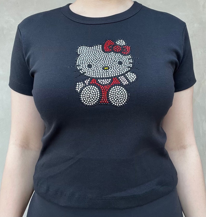 May include: Black t-shirt featuring a rhinestone Hello Kitty design. The design has a white outline, red bow, and black eyes.