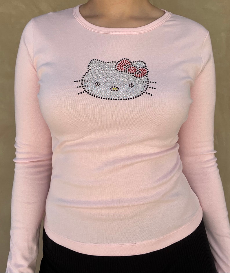 May include: A pink long-sleeved shirt with a rhinestone Hello Kitty design. The design features a white Hello Kitty face with a red bow.