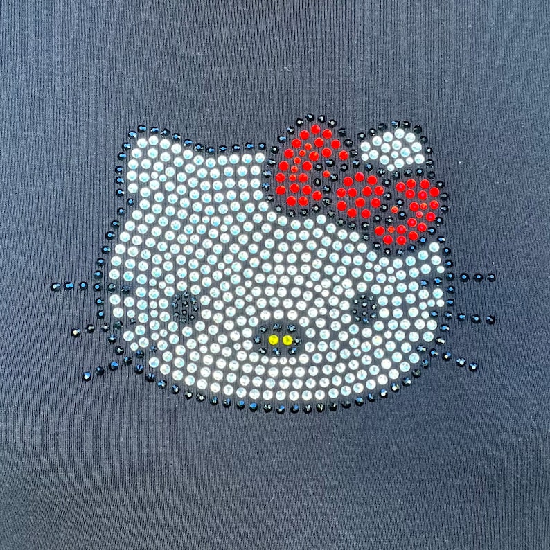 May include: A black t-shirt with a white Hello Kitty design made of rhinestones. The bow on Hello Kitty's head is red and white.