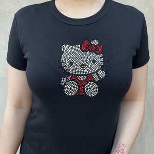 May include: Black t-shirt featuring a rhinestone Hello Kitty design. The design shows Hello Kitty wearing a red dress with a pink bow.