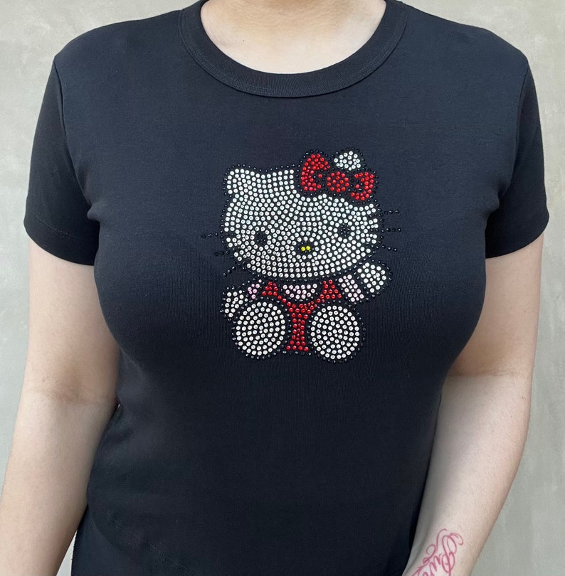 May include: Black t-shirt featuring a rhinestone Hello Kitty design. The design shows Hello Kitty wearing a red dress with a pink bow.
