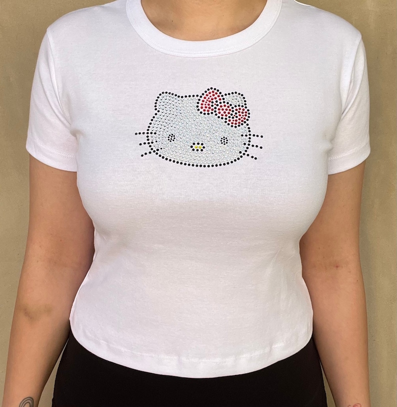 Pu&ograve; includere: Maglietta bianca corta con un design Hello Kitty in strass. Il design include un fiocco rosso e baffi neri.