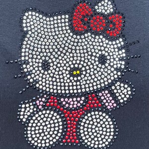 May include: Black t-shirt featuring a rhinestone Hello Kitty design. The design depicts a white Hello Kitty with a red bow and a red bow tie.
