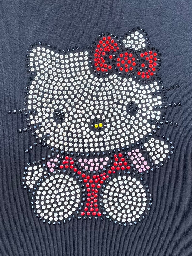 May include: Black t-shirt featuring a rhinestone Hello Kitty design. The design depicts a white Hello Kitty with a red bow and a red bow tie.