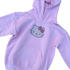 Puede incluir: Una sudadera con capucha de color rosa claro con un dise&ntilde;o de Hello Kitty de diamantes de imitaci&oacute;n en la parte delantera. La sudadera tiene una capucha con cord&oacute;n y un bolsillo canguro.