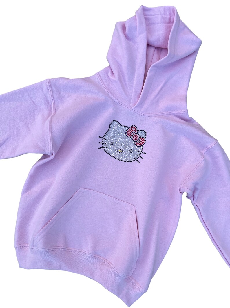 Puede incluir: Una sudadera con capucha de color rosa claro con un dise&ntilde;o de Hello Kitty de diamantes de imitaci&oacute;n en la parte delantera. La sudadera tiene una capucha con cord&oacute;n y un bolsillo canguro.